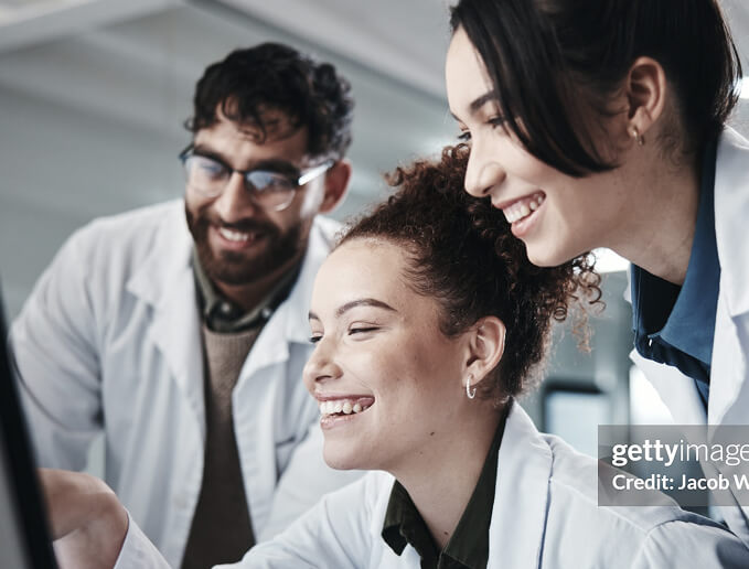 Group of researchers in lab coats smiling while working together at a computer